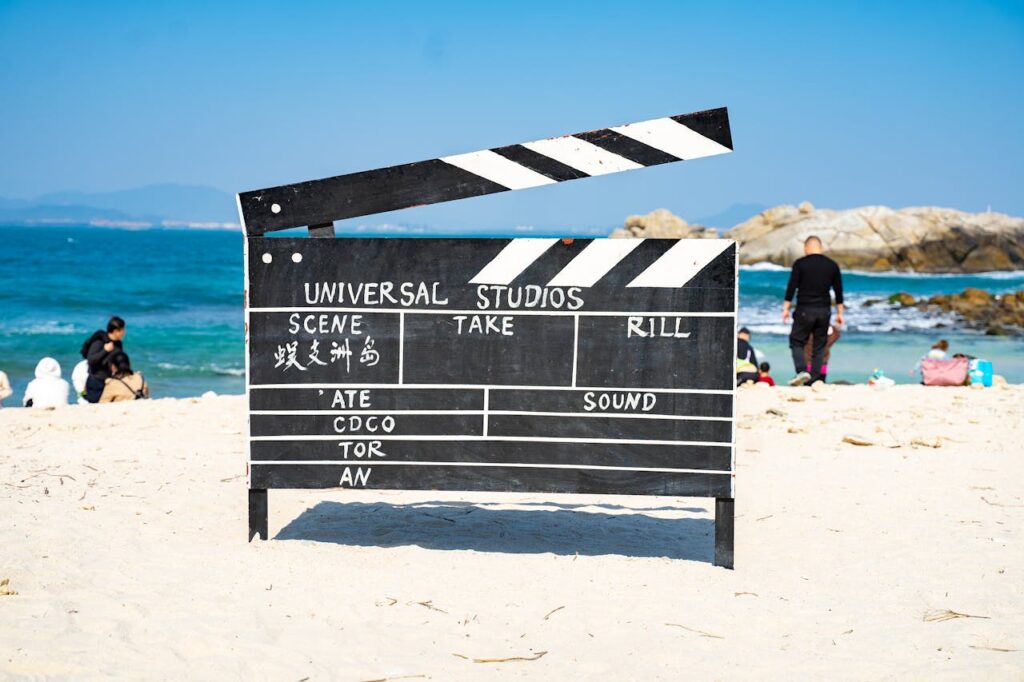 Large film clapperboard on a sandy beach with people and the ocean in the background.
